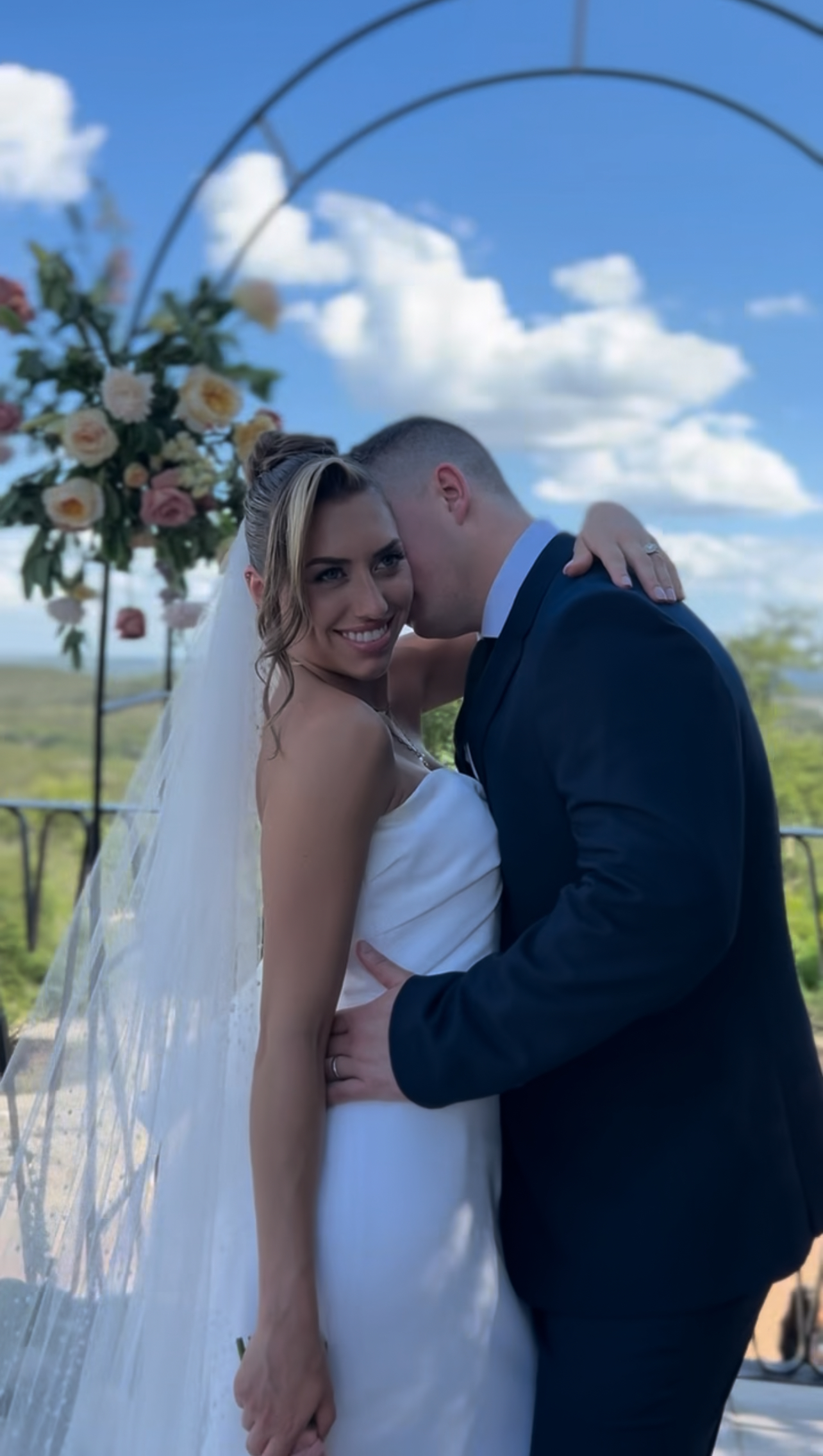 A bride and groom embrace under a floral arch outdoors, with a bright blue sky and white clouds in the background.