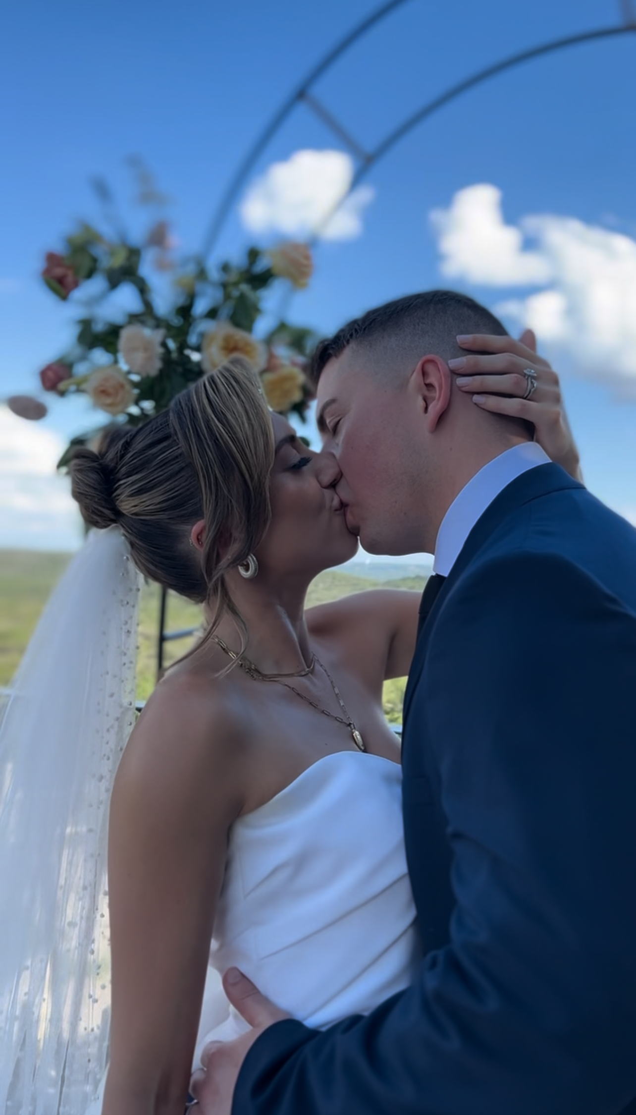 A bride and groom embrace under a floral arch outdoors, with a bright blue sky and white clouds in the background.