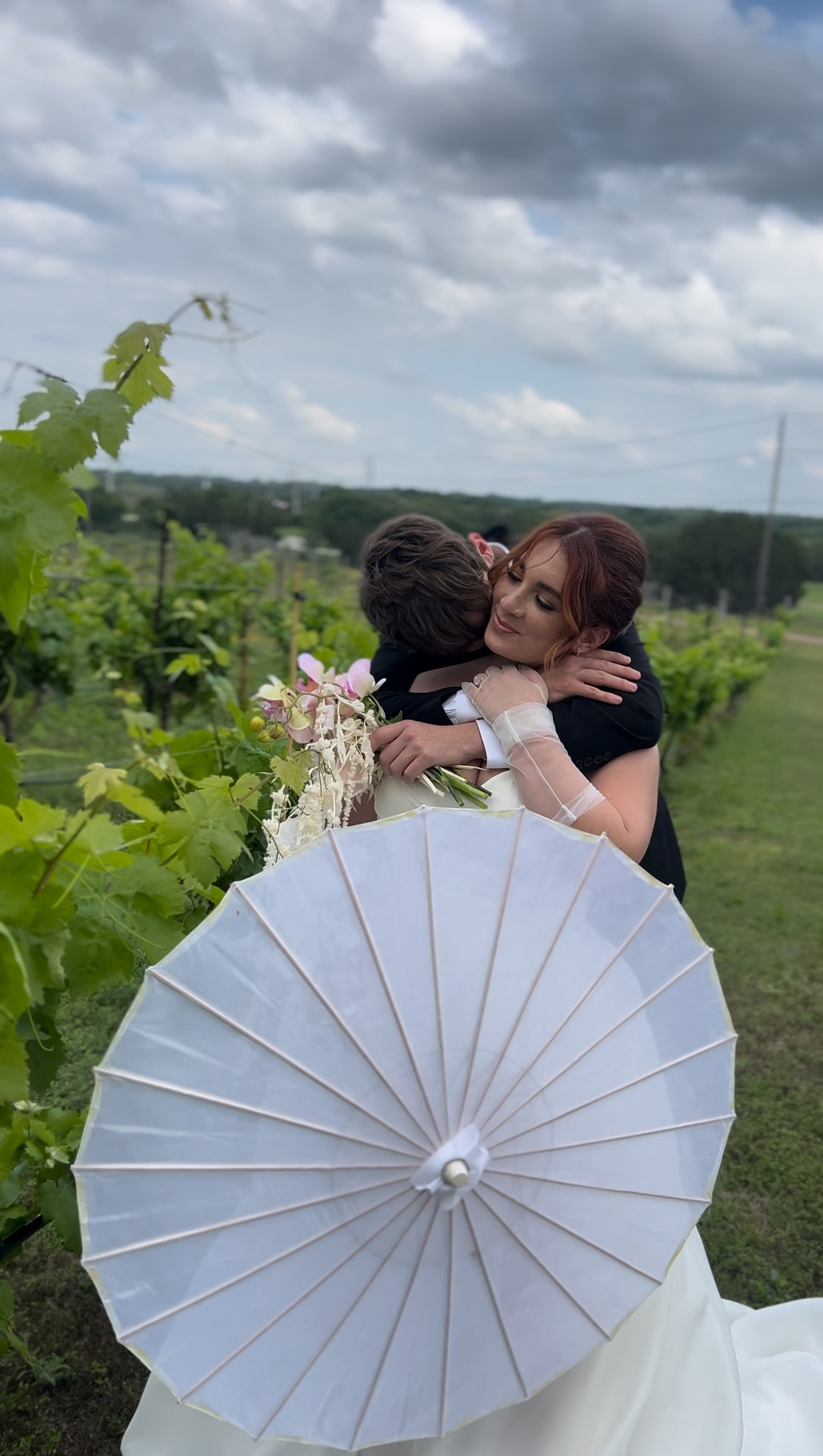 A bride and groom embrace in a vineyard, with the bride holding a white parasol and bouquet. Cloudy sky and green vines are in the background.