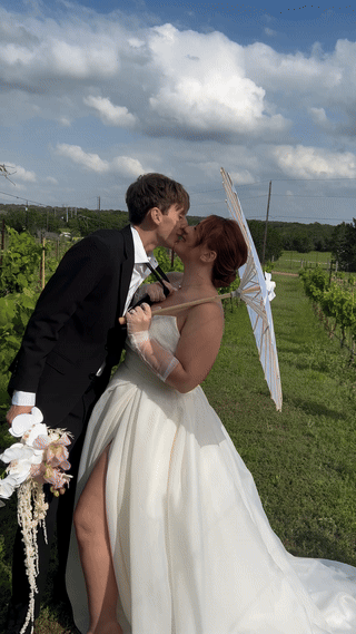 A bride and groom share a kiss outdoors. The bride holds a parasol and bouquet, while the groom wears a suit. They stand on a grassy field under a partly cloudy sky captured by Austin wedding content creator 