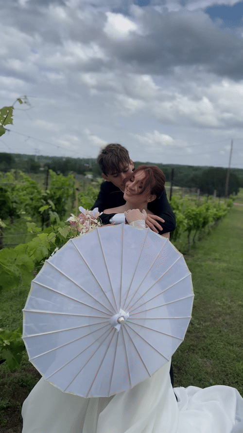A bride and groom share a kiss outdoors. The bride holds a parasol and bouquet, while the groom wears a suit. They stand on a grassy field under a partly cloudy sky captured by Austin wedding content creator