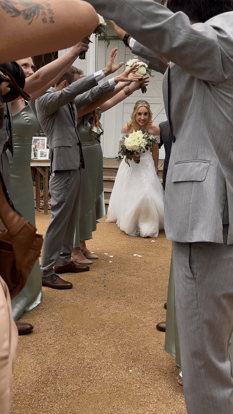 Bride walking under an archway formed by people in gray suits and green dresses, holding bouquets.