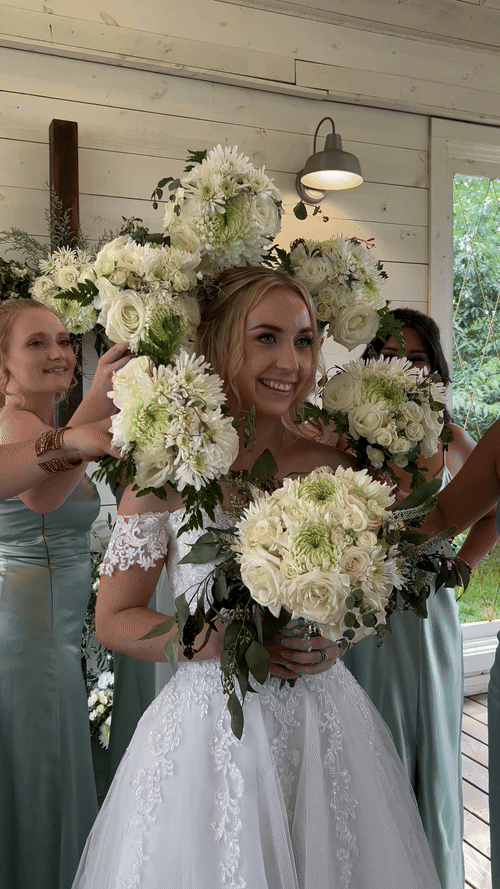 Bride smiling with bridesmaids placing a bouquet of white flowers around her head. She holds a matching bouquet in front. Bridesmaids wear light green dresses captured by an austin wedding content creator
