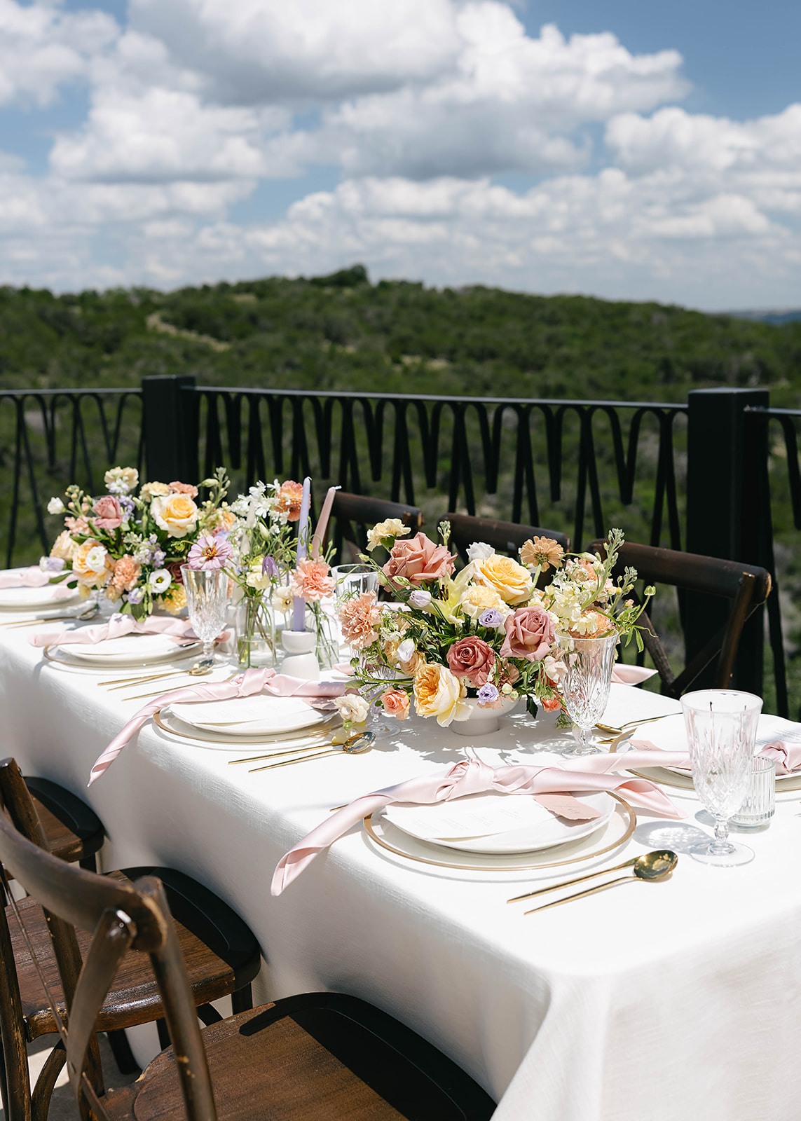Elegant outdoor dining table with floral centerpieces, white tablecloth, and place settings. Scenic view of lush green hills under a partly cloudy sky in the background at wedding venue Villa Antonia 