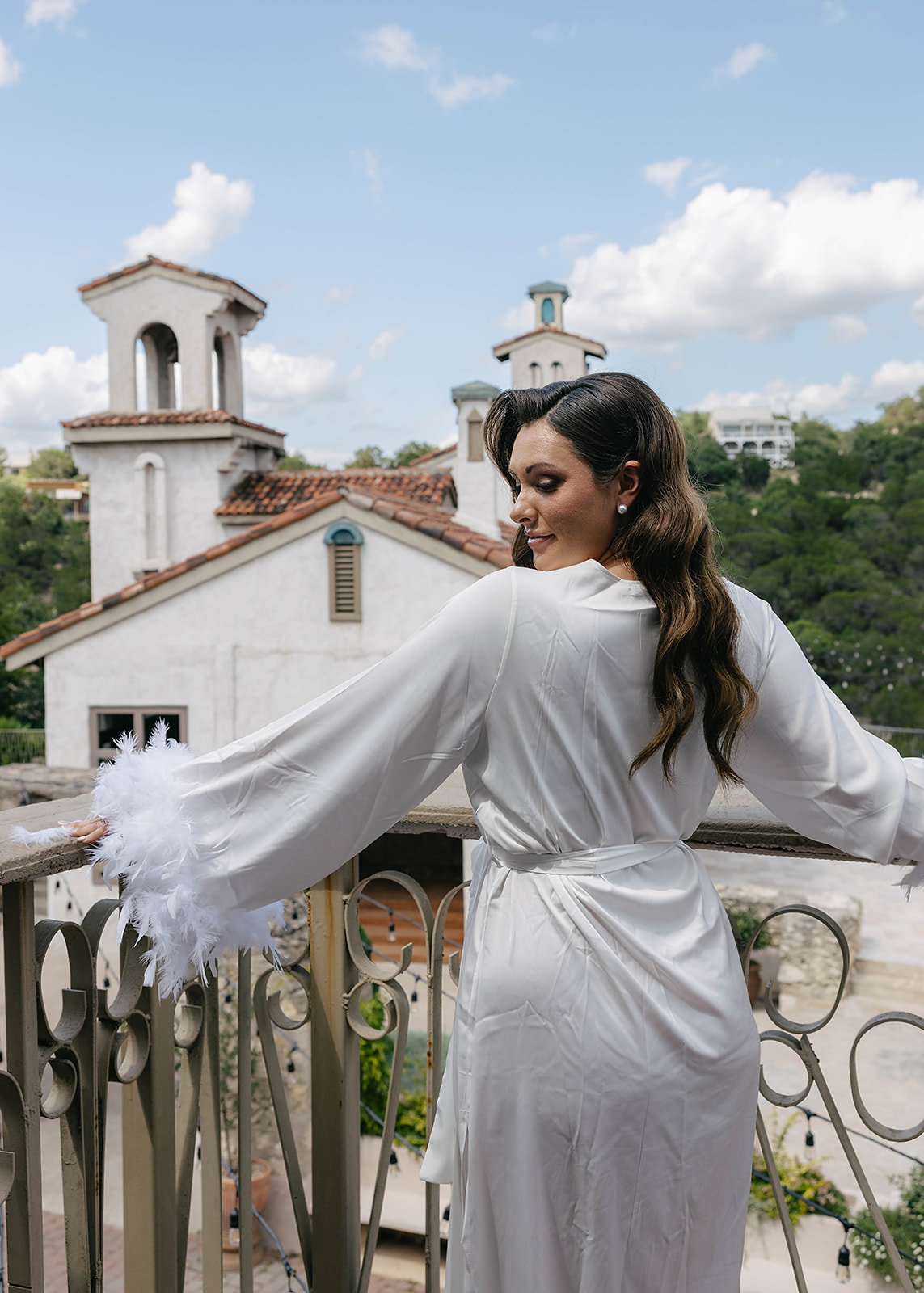 A woman in a white robe stands on a balcony overlooking green trees and white buildings under a partly cloudy sky at Villa antonia