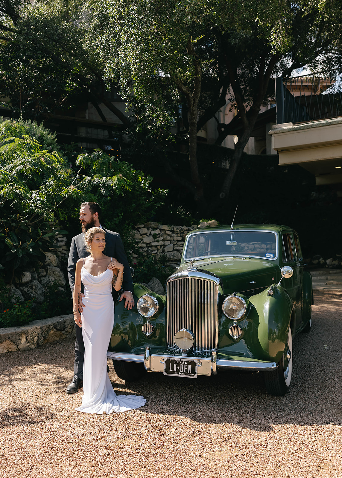 A couple stands next to a vintage green car in an outdoor setting with trees and a building in the background.