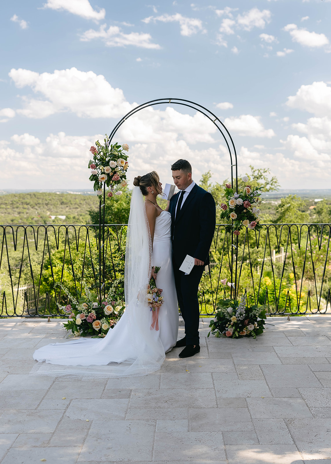 Bride and groom stand under a floral arch outdoors, with a scenic view in the background at Villa antonia