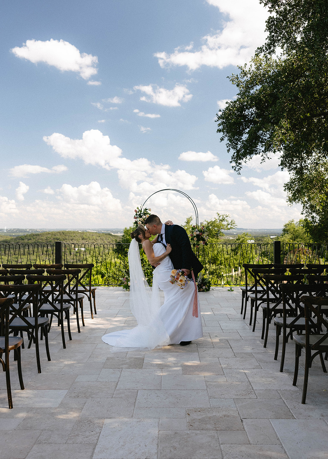 Bride and groom stand under a floral arch outdoors, with a scenic view in the background at Villa antonia