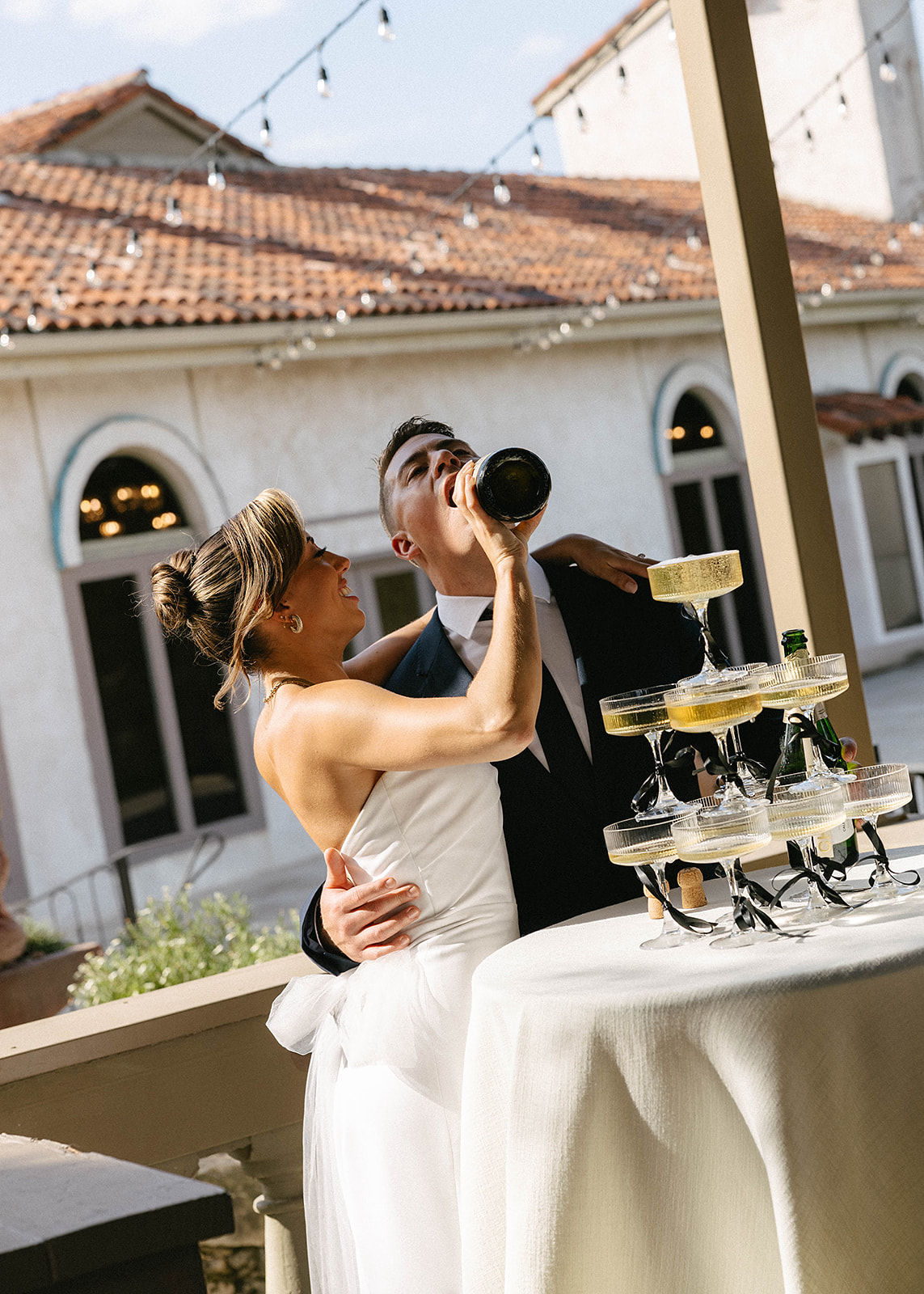 A bride and groom pour champagne together onto a pyramid of glasses at a reception, set in a venue with Mediterranean-style architecture.