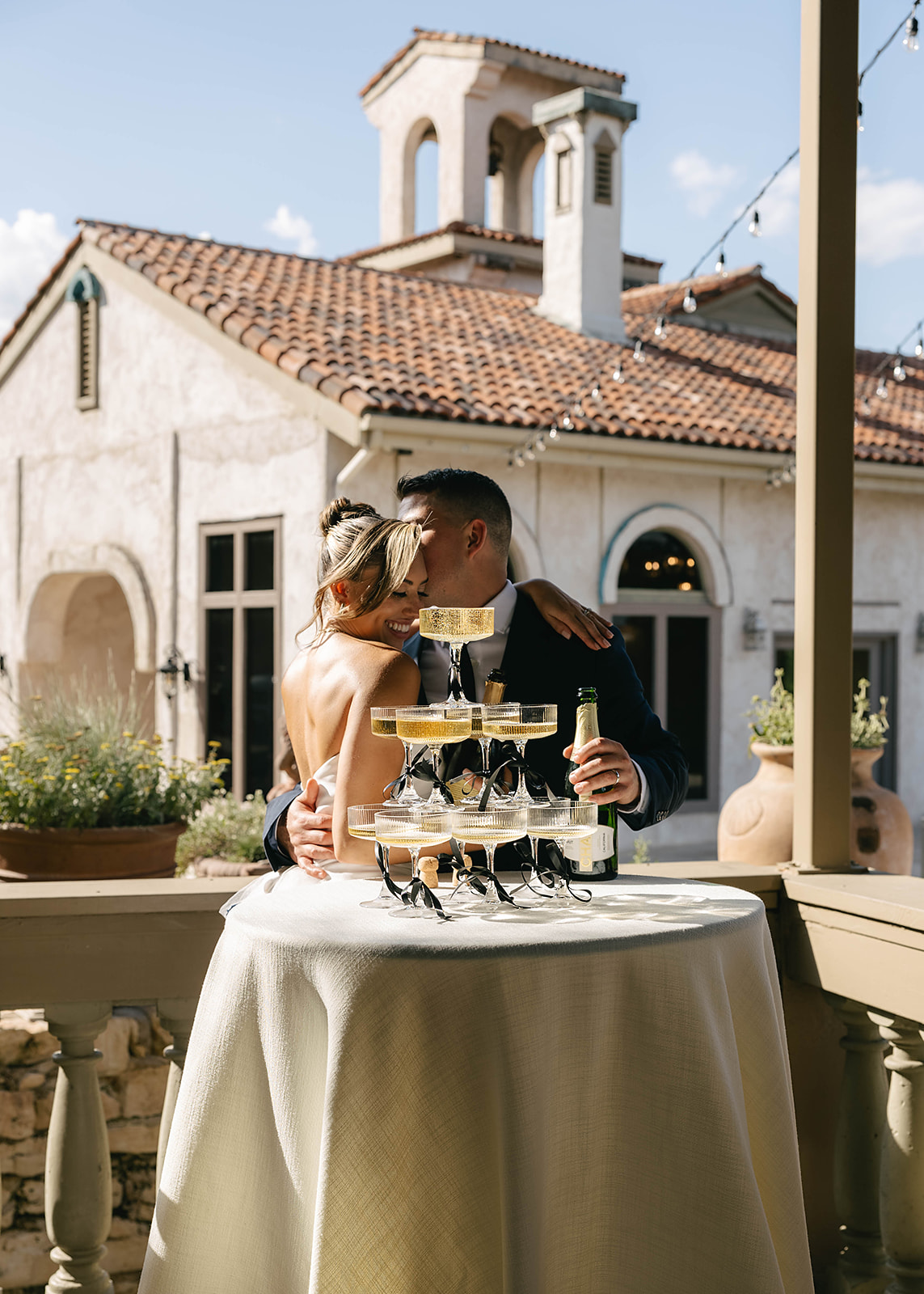 A bride and groom pour champagne together onto a pyramid of glasses at a reception, set in a venue with Mediterranean-style architecture.